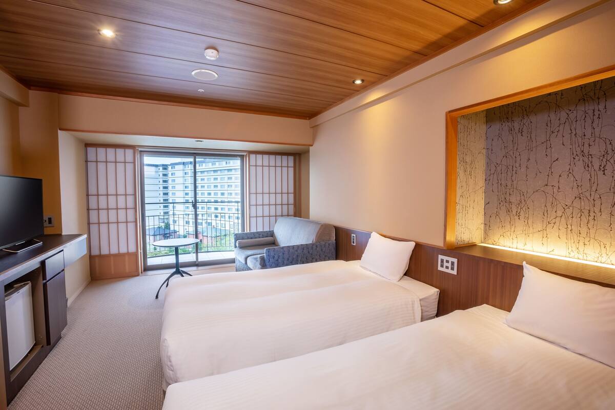 A hotel room with two beds, a TV, a desk, and a view of a building through a large window.