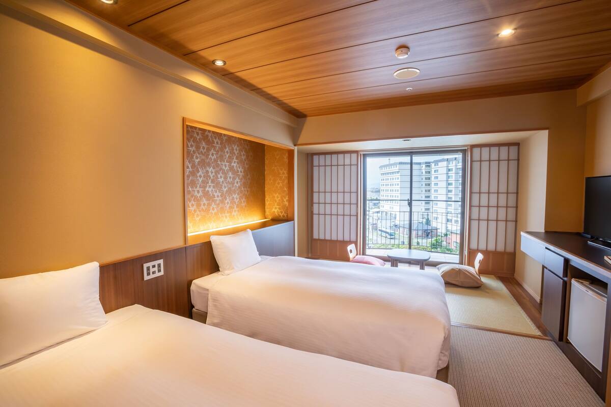 A hotel room with two beds, a TV, a desk, and a view of a building through a window.