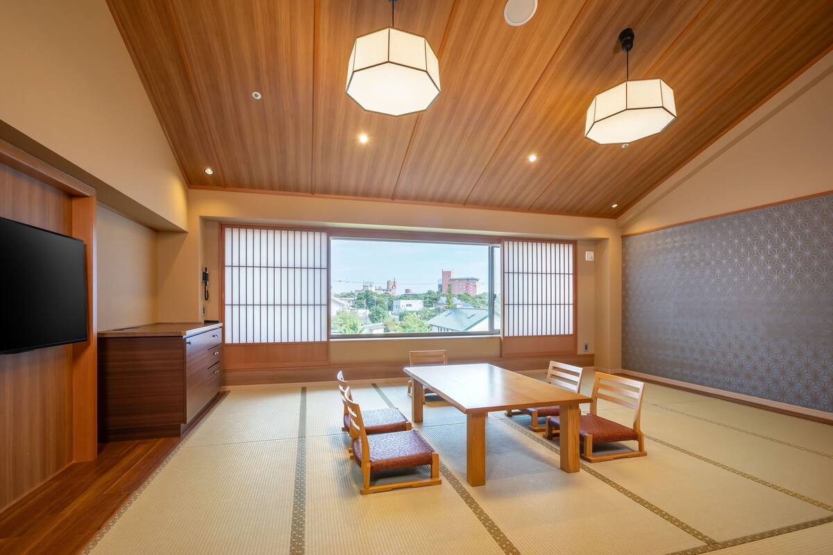 A traditional Japanese-style room with tatami flooring, a wooden ceiling, and a large window offering a city view.