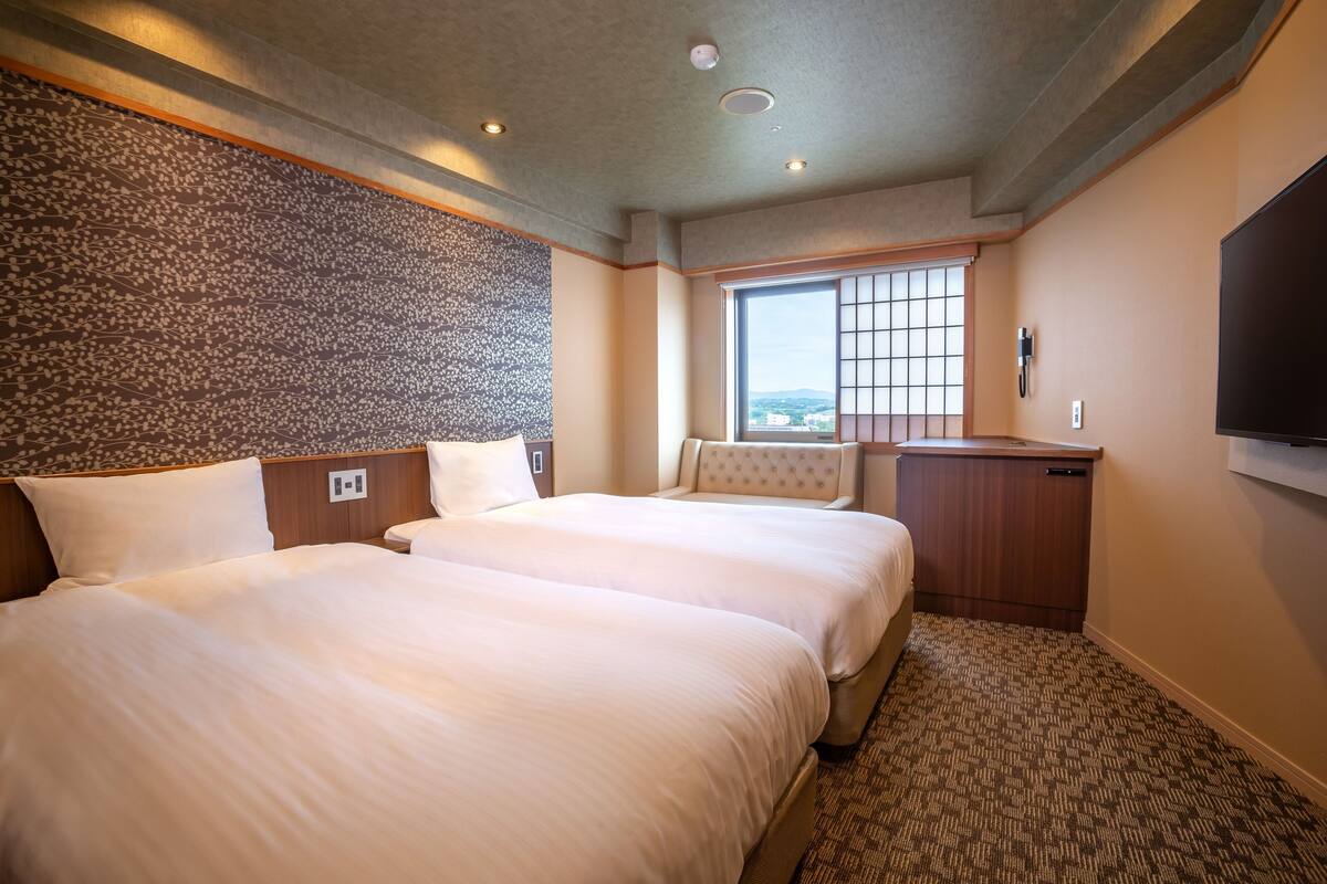 A hotel room with two beds, a window with a city view, a flat-screen TV, and a small desk area.