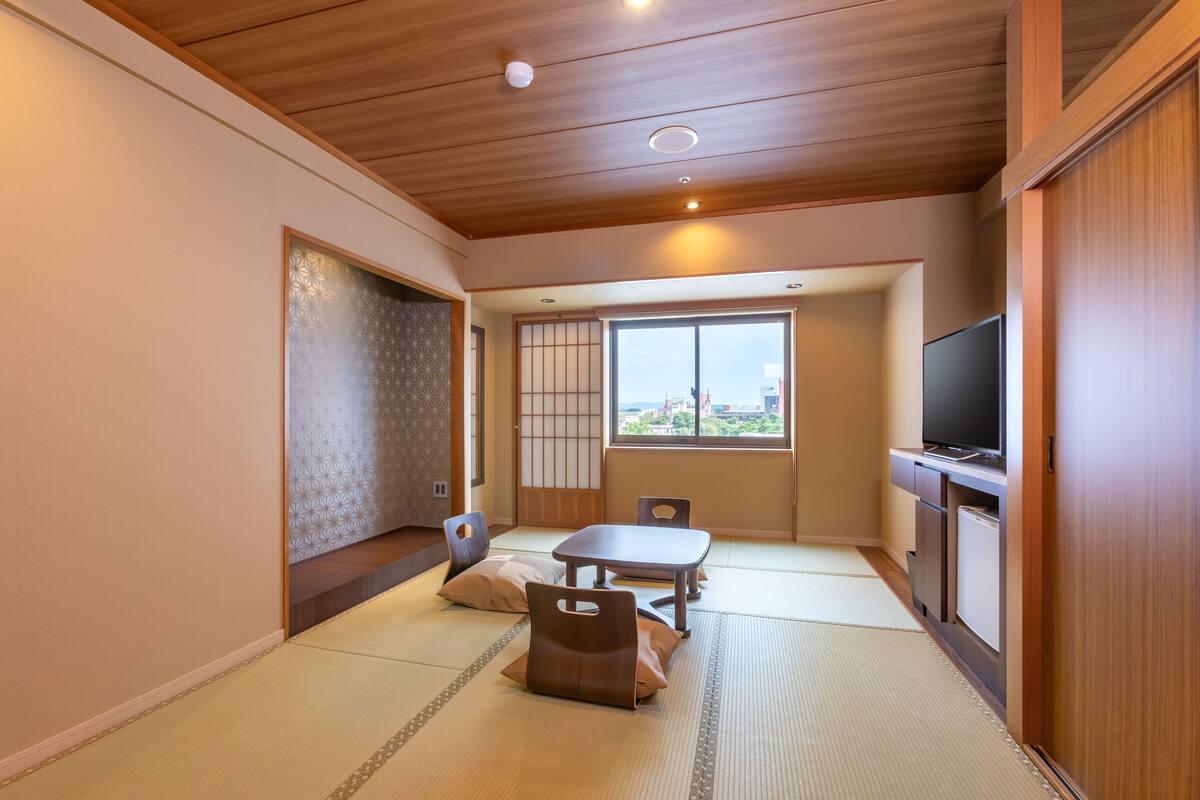 A traditional Japanese room with tatami flooring, a low table, cushions, a television, and a sliding door leading to a balcony.