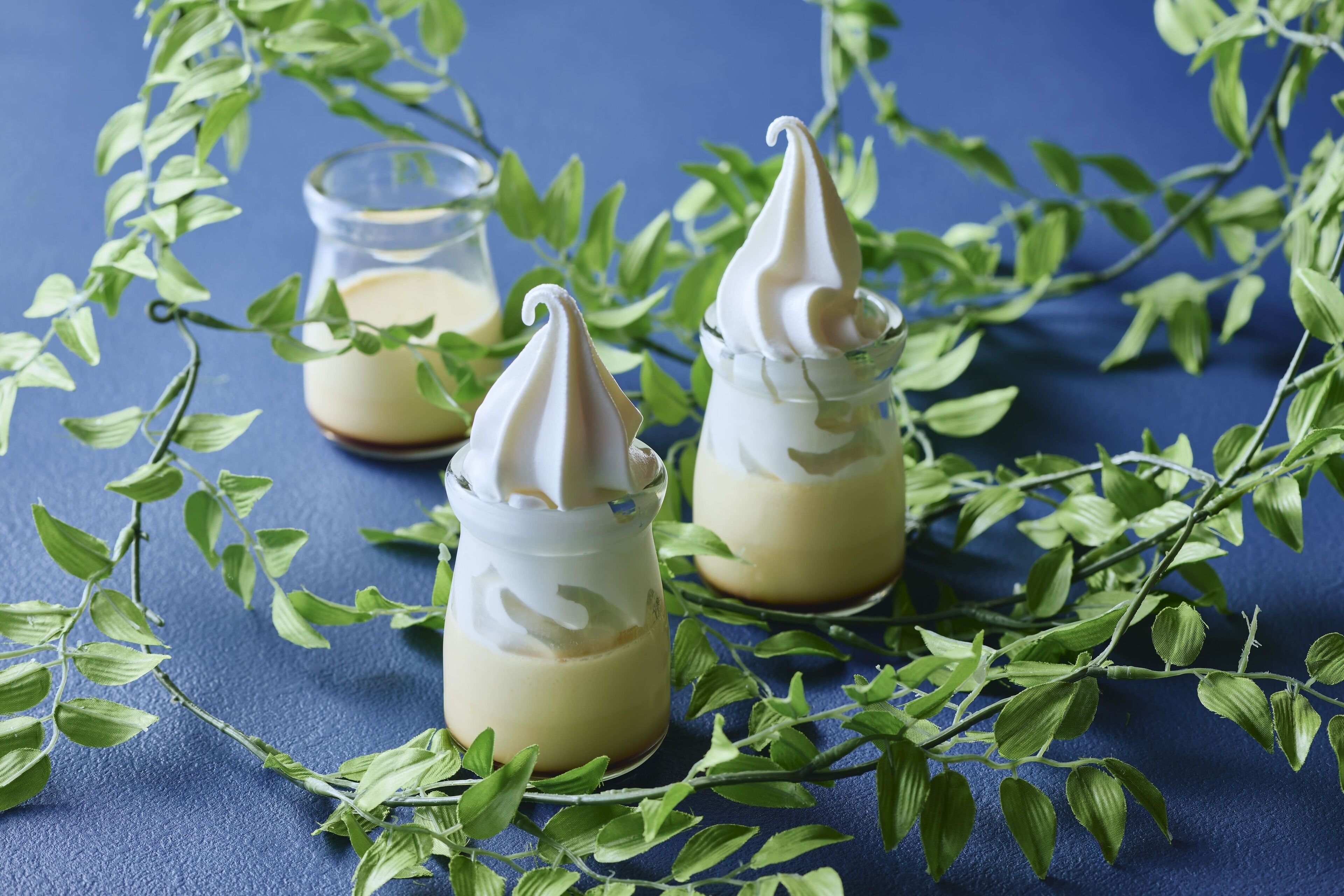 Three glass jars with a creamy substance and a whipped topping, surrounded by green leaves on a blue background.