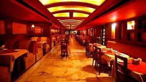 A long, narrow hotel corridor with tables and chairs on both sides, leading to a bright, open area.