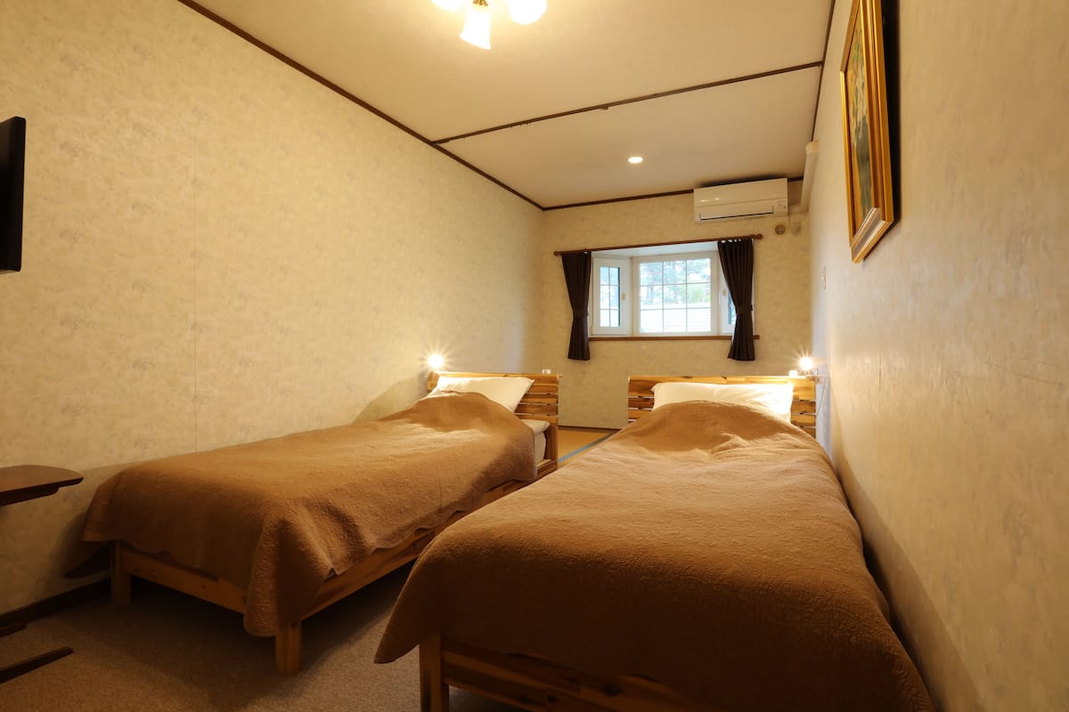 A room with two wooden beds, a small table, a window with curtains, and an air conditioning unit on the wall.