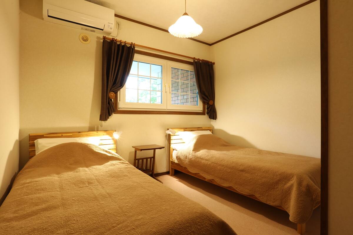 A room with two single beds, a small table, a window with curtains, and an air conditioning unit.