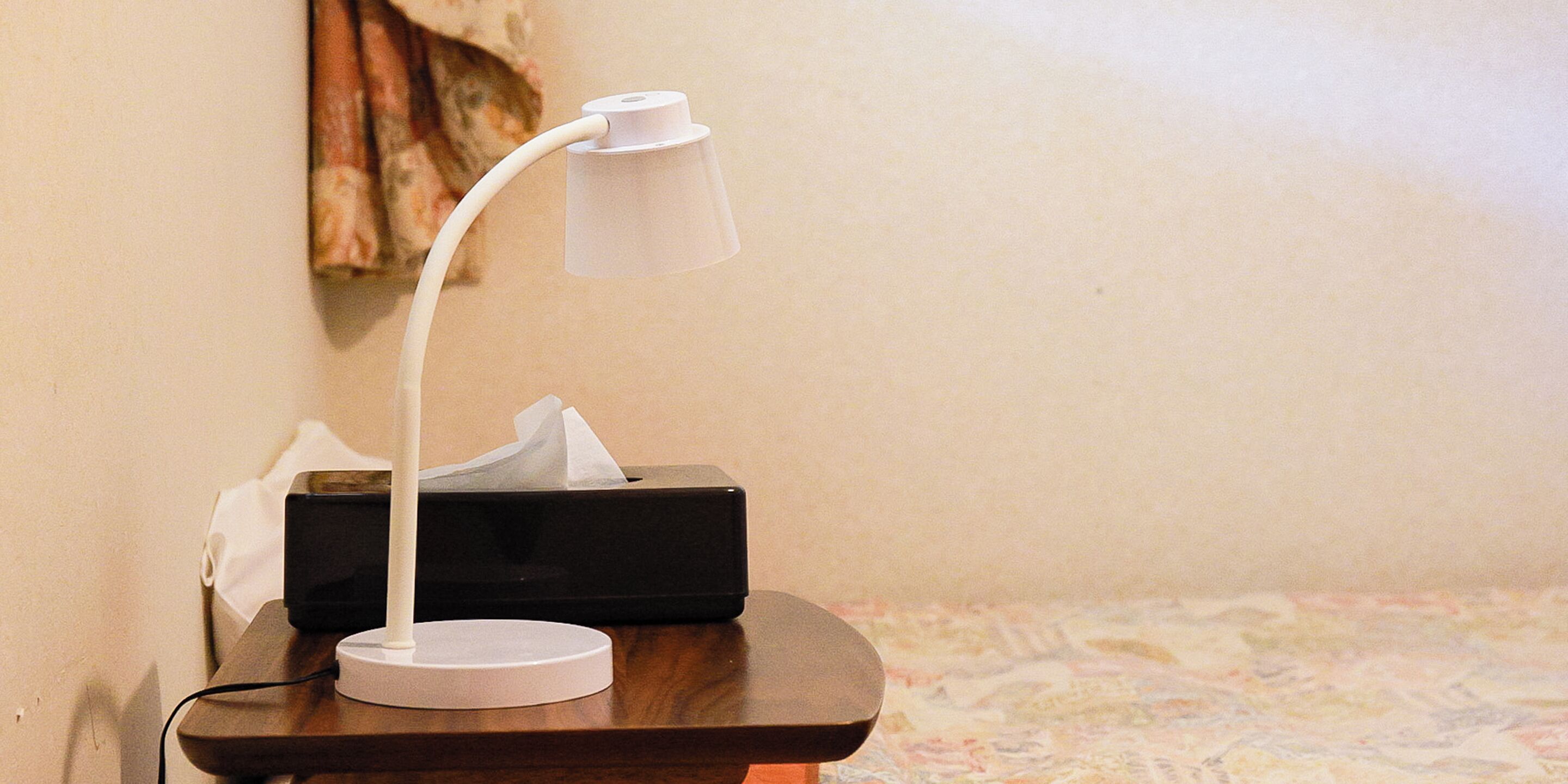 A desk lamp on a wooden table with a tissue box and a black electronic device in the background.