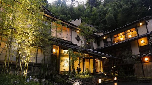 A traditional Japanese-style building with multiple levels, illuminated windows, and a garden area with rocks and plants.