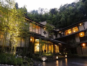 A traditional Japanese-style building with multiple levels, illuminated windows, and a garden area with rocks and plants. - Yuraku Kinosaki Spa & Gardens (Toyooka)