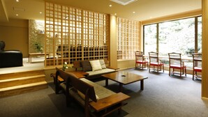 A modern living room with a sofa, coffee table, and chairs. There is a wooden partition with a grid pattern, a staircase, and a large window with a view of greenery.