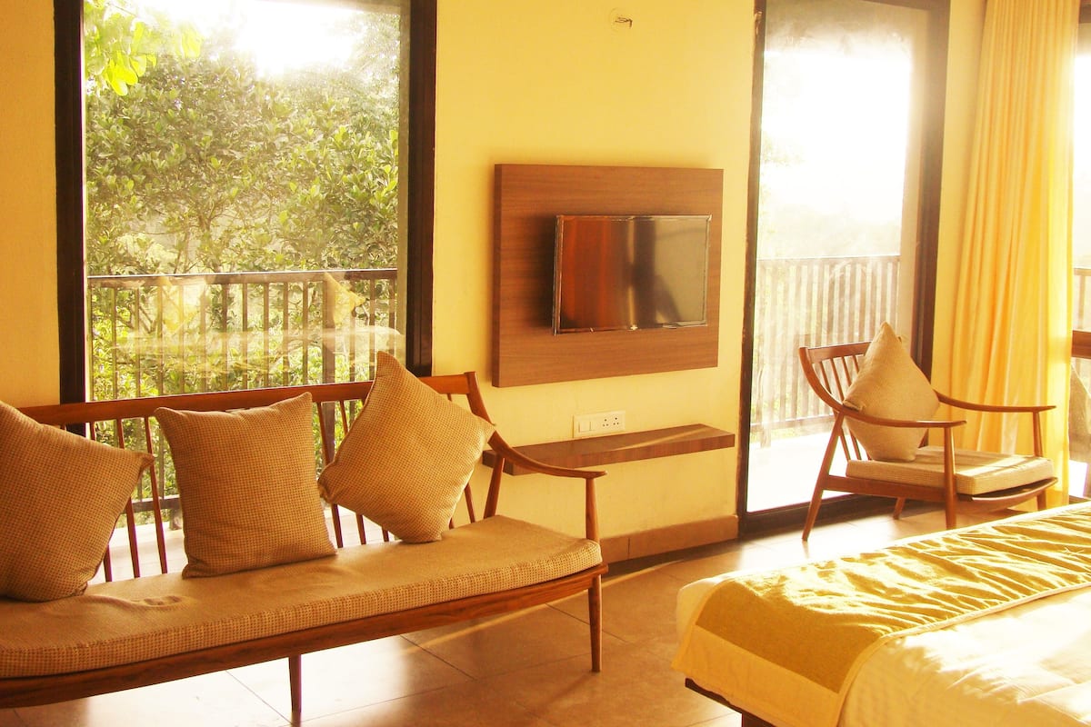 A hotel room with a sofa, a bed, a TV mounted on the wall, and a balcony with a view of greenery.