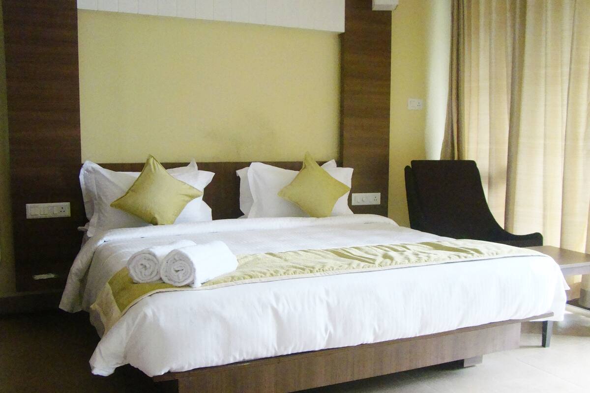 A neatly made bed with white linens and rolled towels, a wooden headboard, and a black chair to the side.