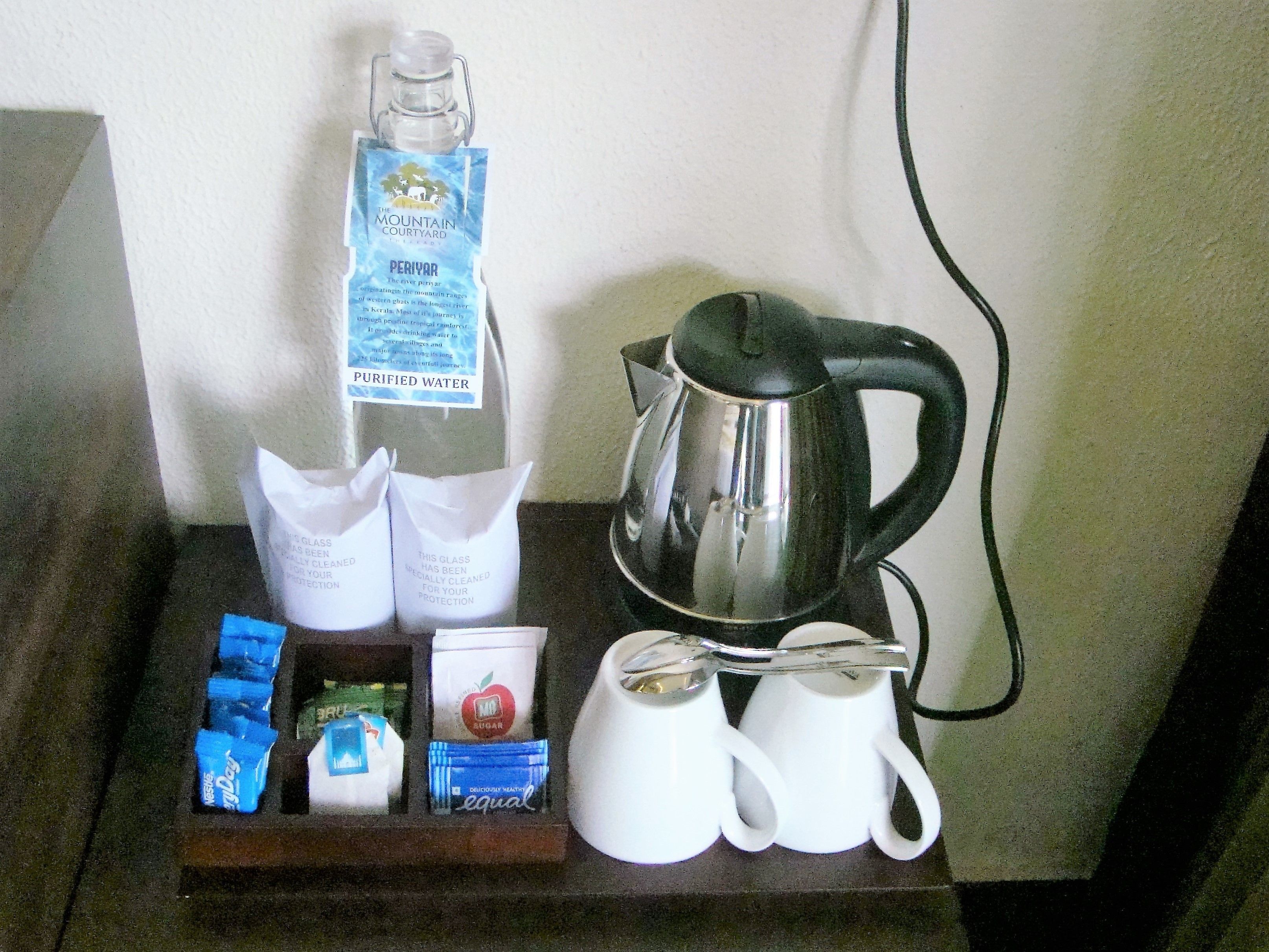 Un plateau avec un verre d’eau purifiée, une bouilloire, des tasses blanches et des sachets de sucre et de thé.