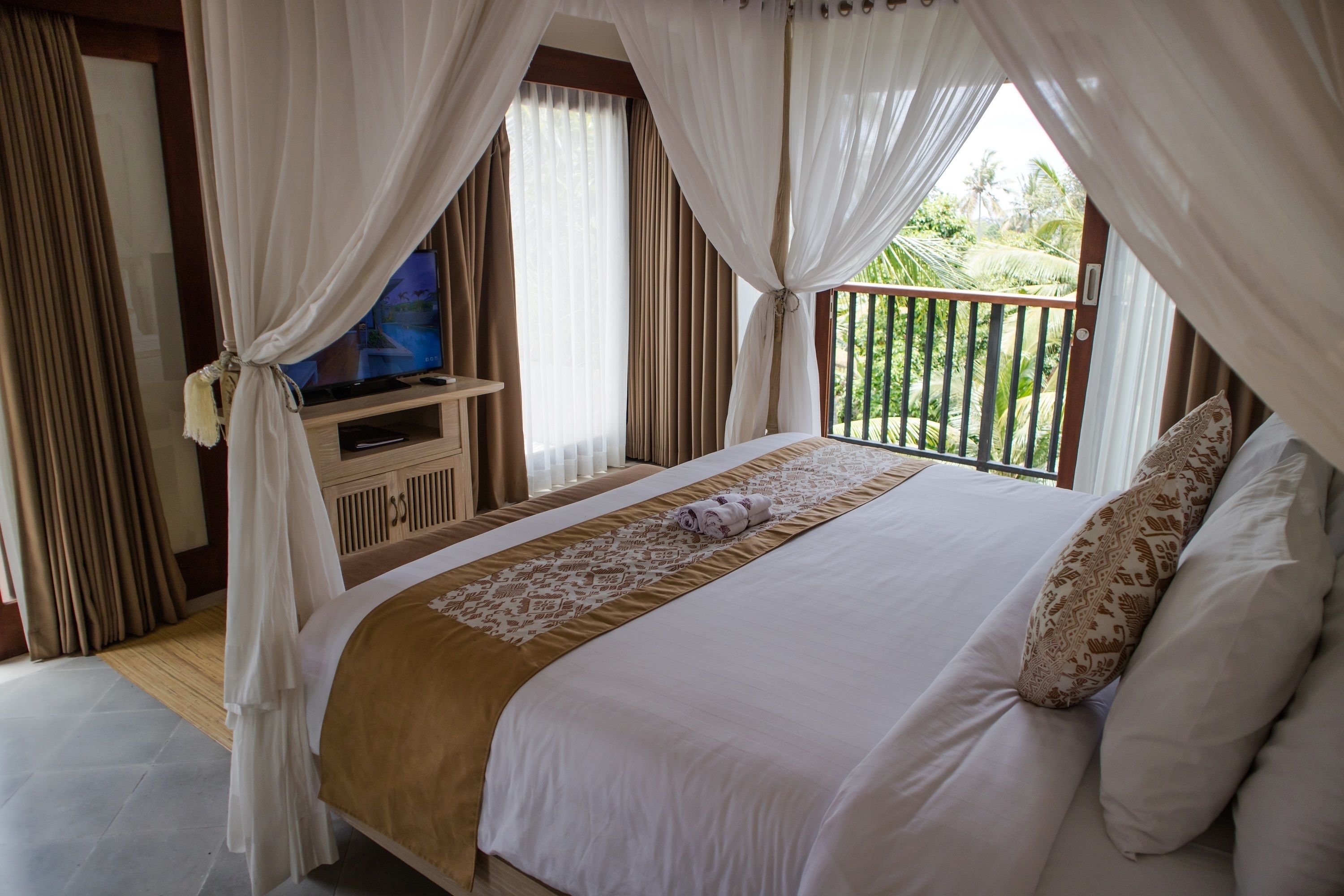 A bedroom with a bed, curtains, a television, and a balcony with a view of greenery.