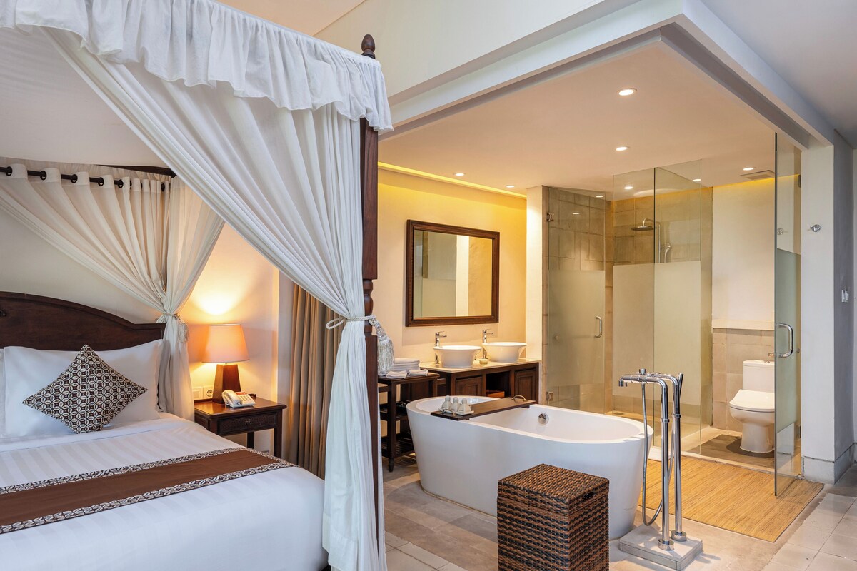 A hotel room with a large bed, a sitting area with a lamp, a bathroom with a bathtub, and a glass-enclosed shower area.