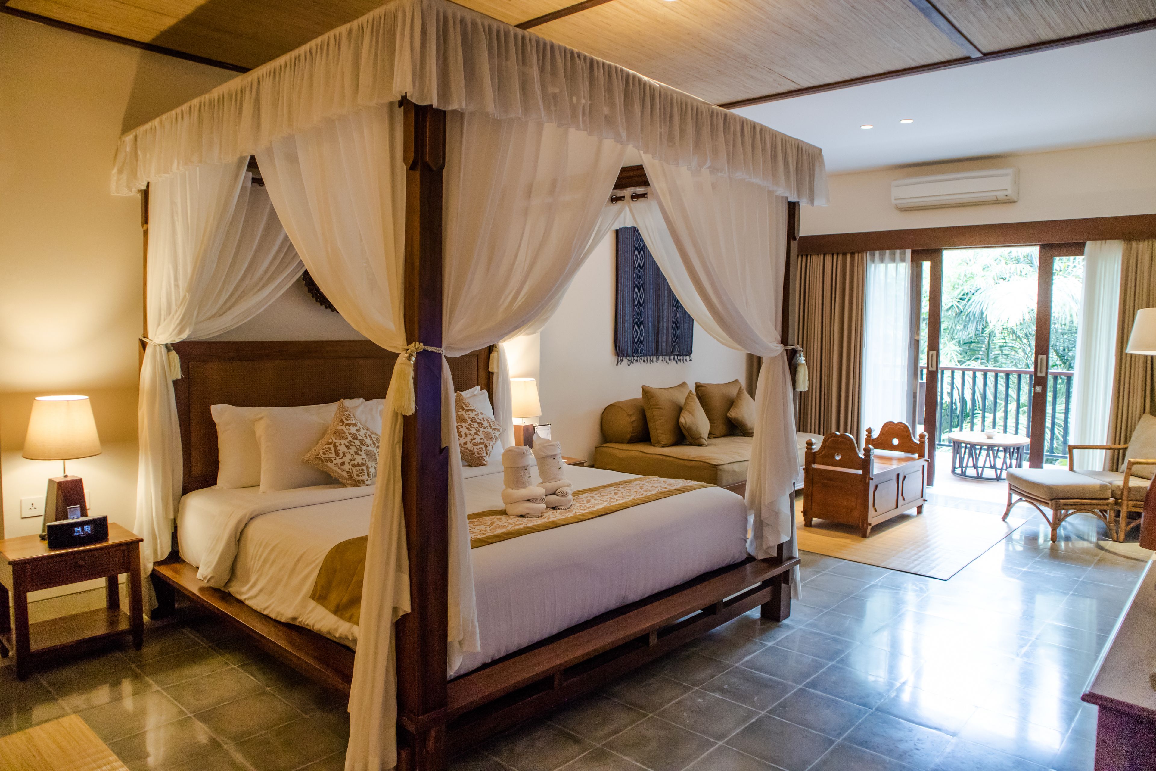 A four-poster canopy bed with a wooden frame, a bedside table with a lamp, and a view of the outdoor balcony.