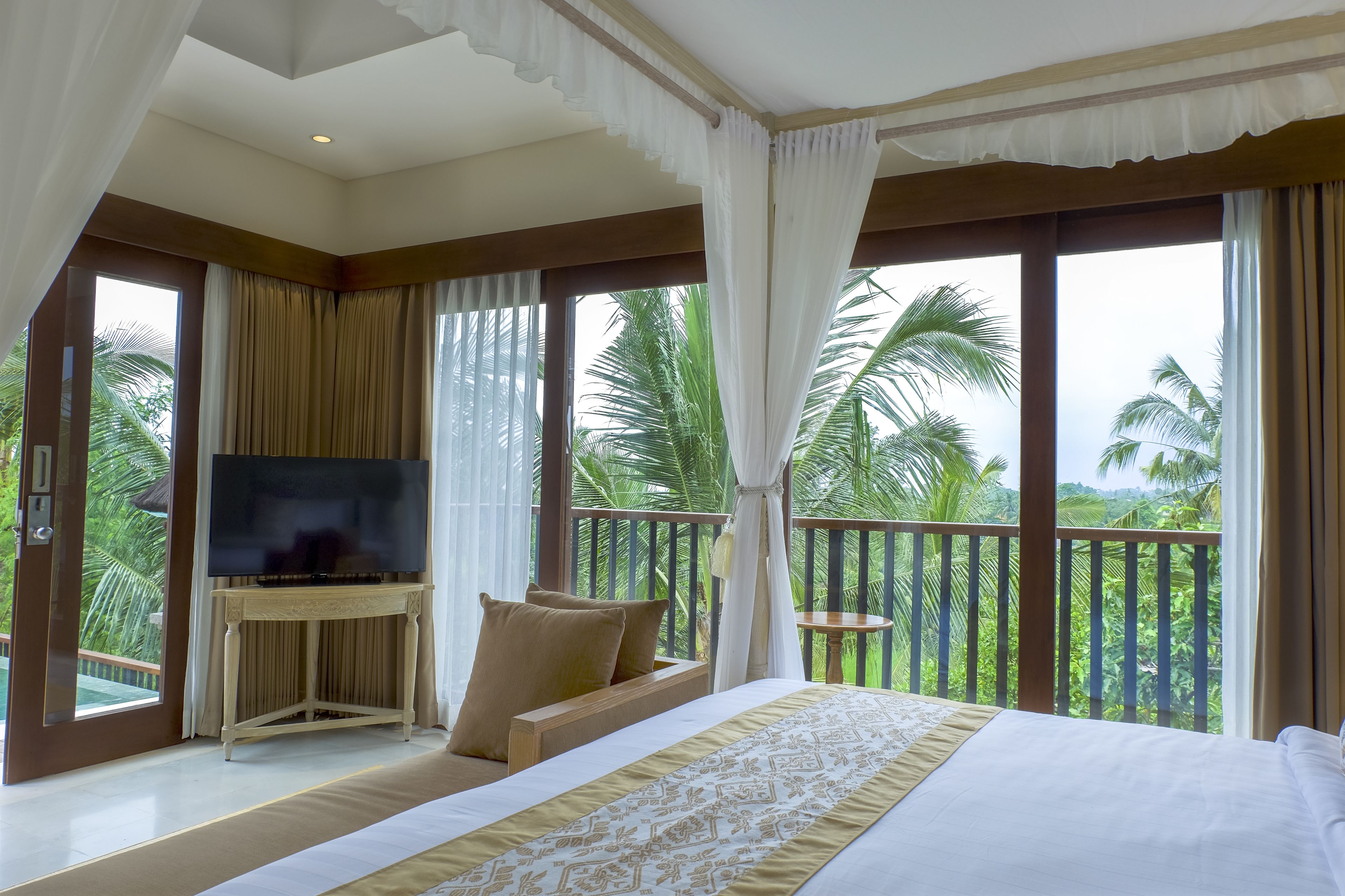 A bedroom with a bed, a TV on a stand, and a balcony with a view of palm trees.