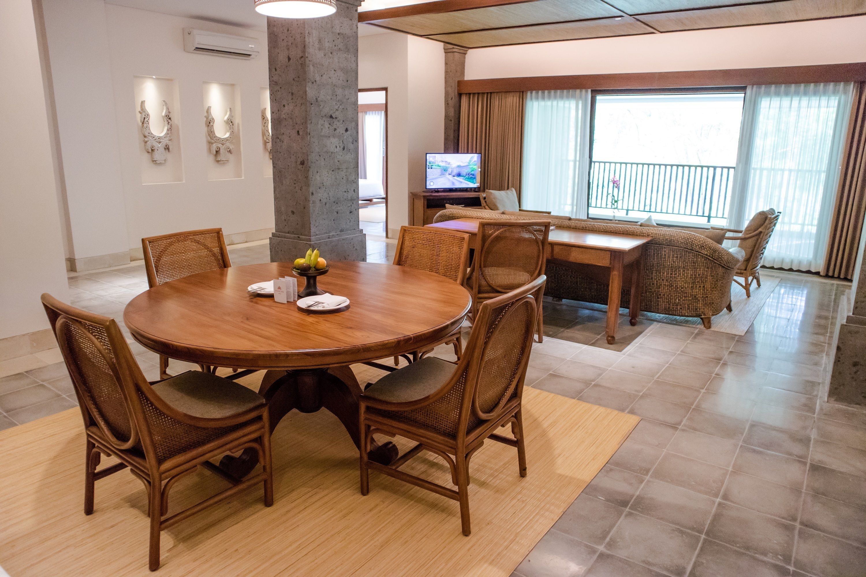 A hotel room with a round dining table, four wicker chairs, a television, and a balcony with curtains.