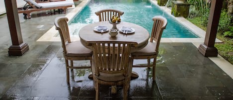 A poolside area with a wooden table and chairs, a covered seating area with lounge chairs, and a lush garden backdrop.