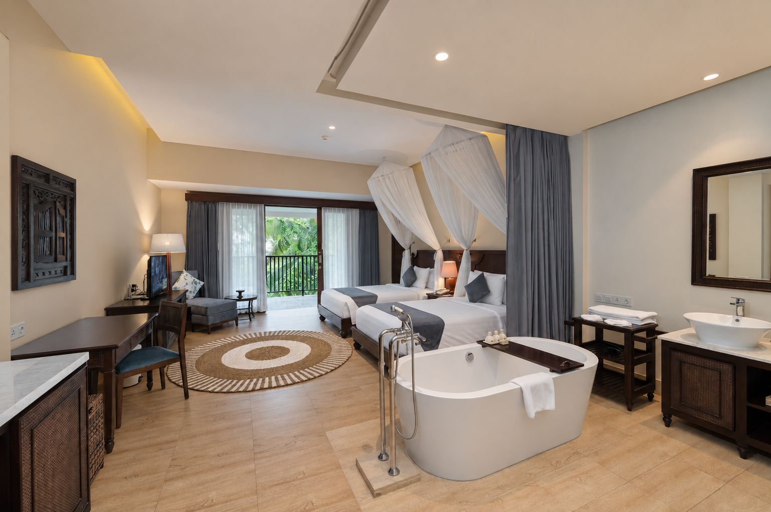 A hotel room with a large bed, a sitting area with a desk and chair, a bathroom with a bathtub and sink, and a balcony with curtains.