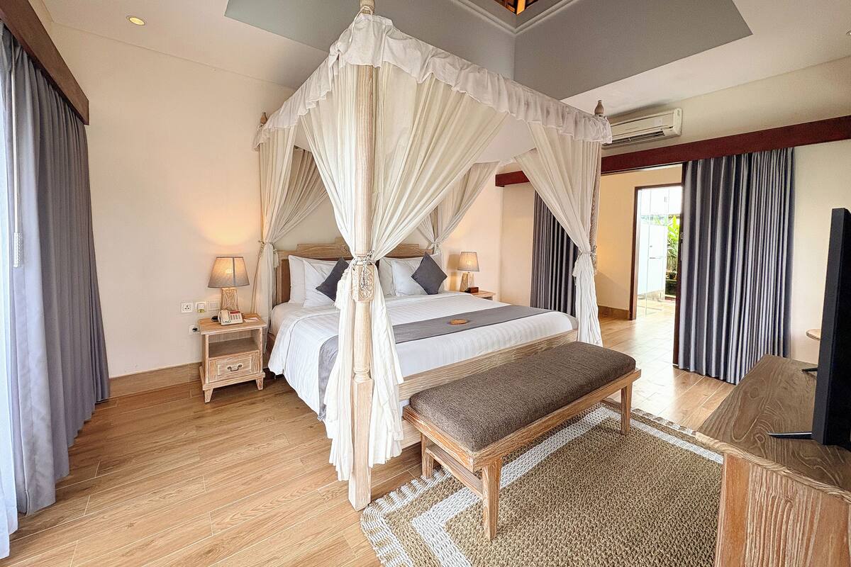 A spacious bedroom with a four-poster canopy bed, a wooden bench, a bedside table with lamps, and a large window with curtains.