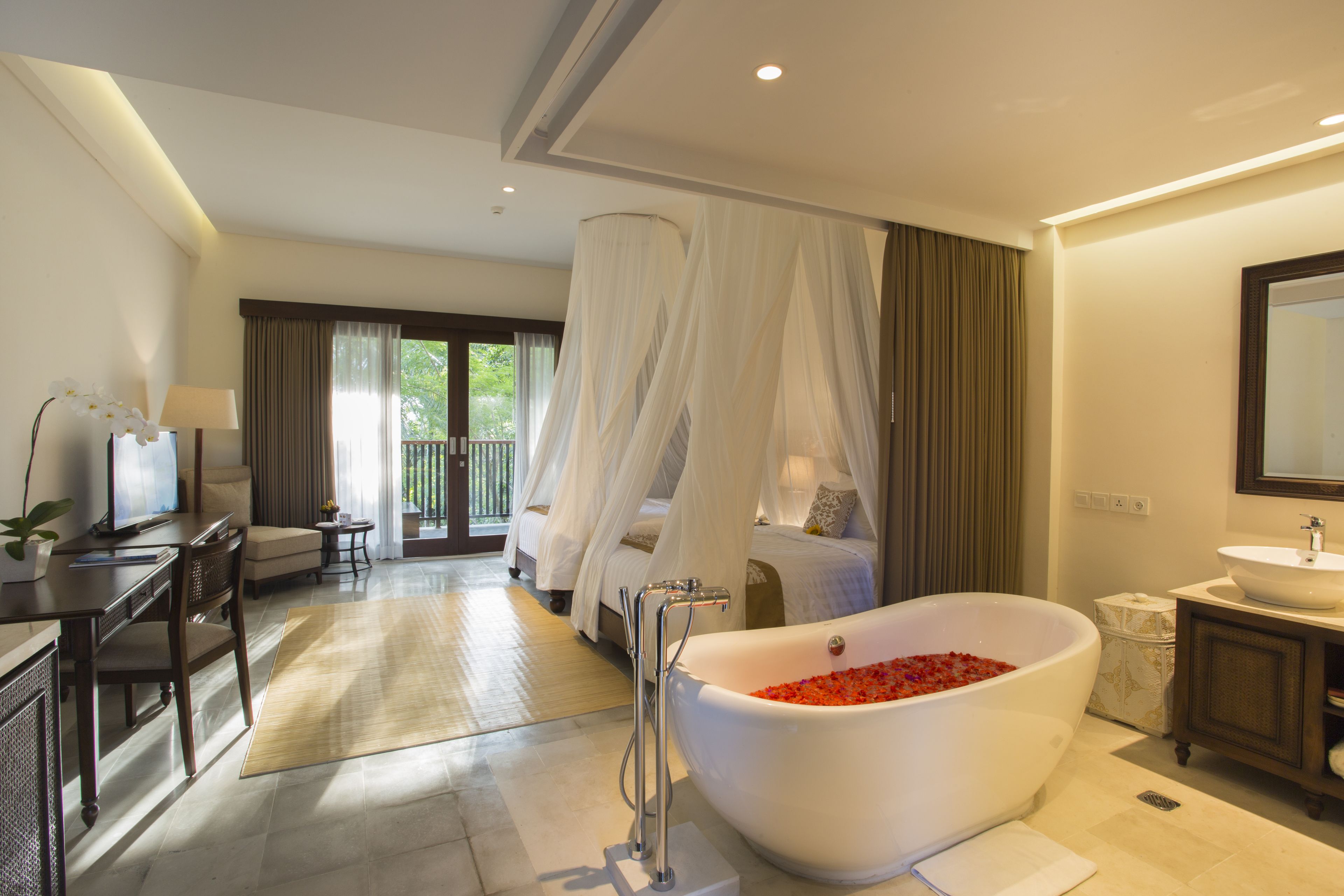 A bedroom with a large bed, a sitting area with a desk and chair, a bathroom with a freestanding bathtub, and a large window with curtains.