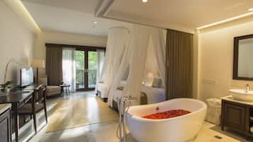 A bedroom with a large bed, a sitting area with a desk and chair, a bathroom with a freestanding bathtub, and a large window with curtains.
