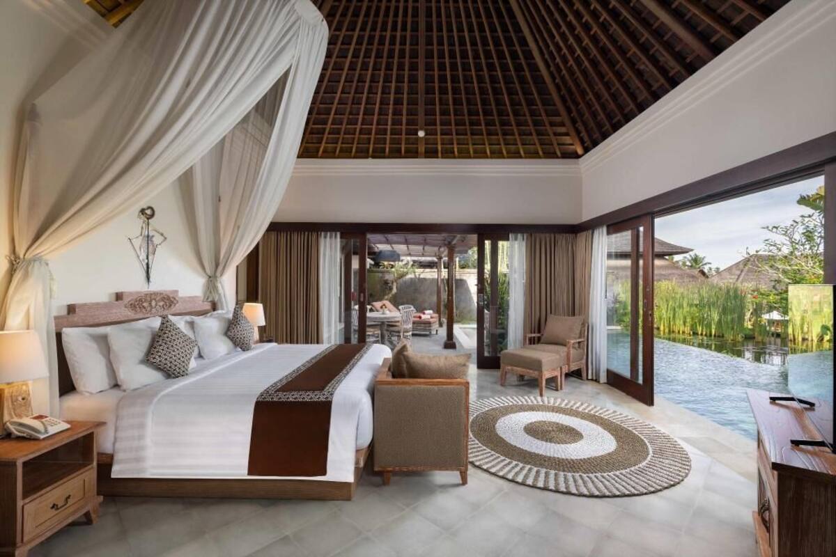 A bedroom with a large bed, a sitting area with chairs, a dining table, and a view of a water body.