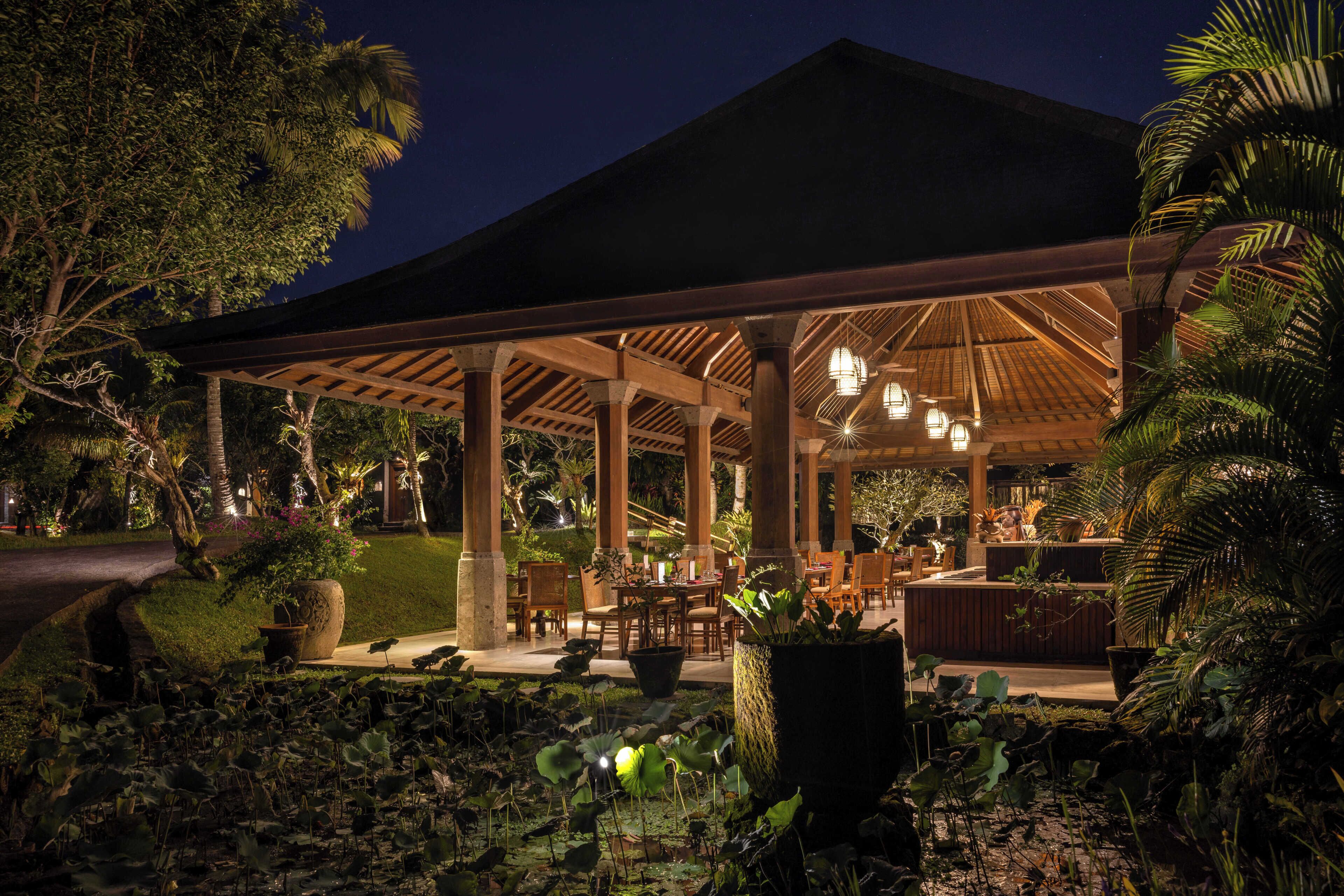 An outdoor covered dining area with wooden pillars, tables, and chairs, surrounded by lush greenery and a pond with lily pads.
