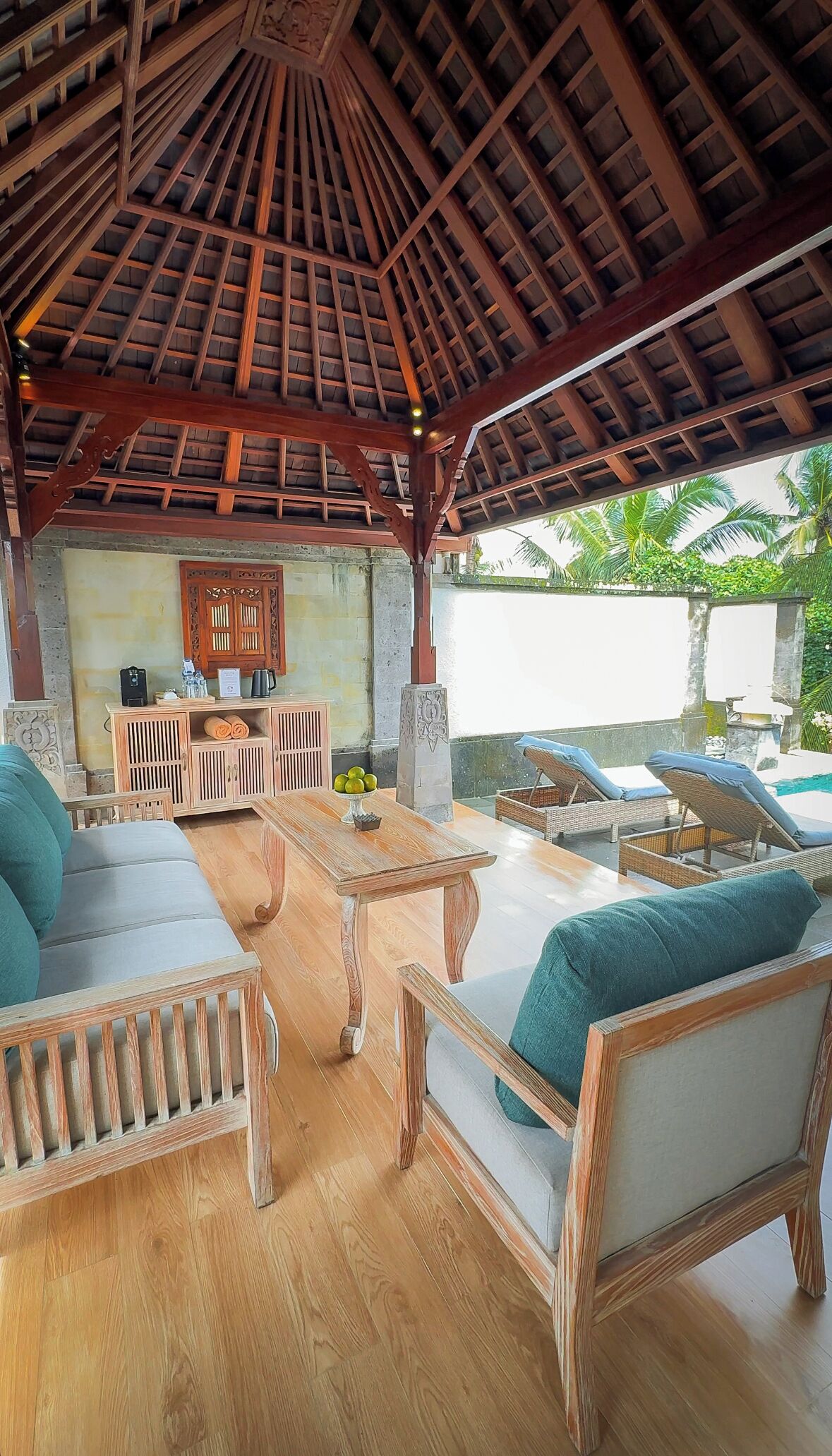 A covered outdoor seating area with wooden furniture, a table, and a view of a swimming pool.