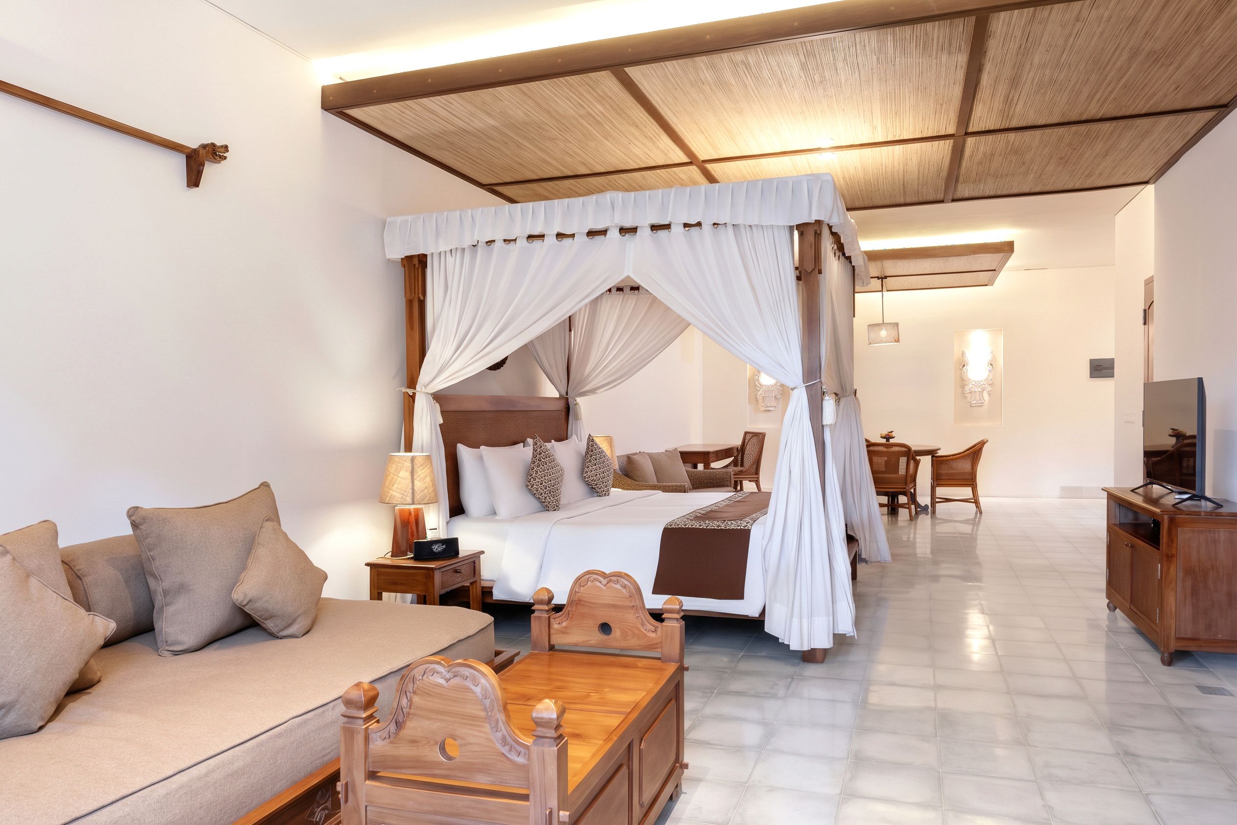 A spacious bedroom with a four-poster canopy bed, a sitting area with a sofa and a wooden table, and a dining area with wicker chairs.