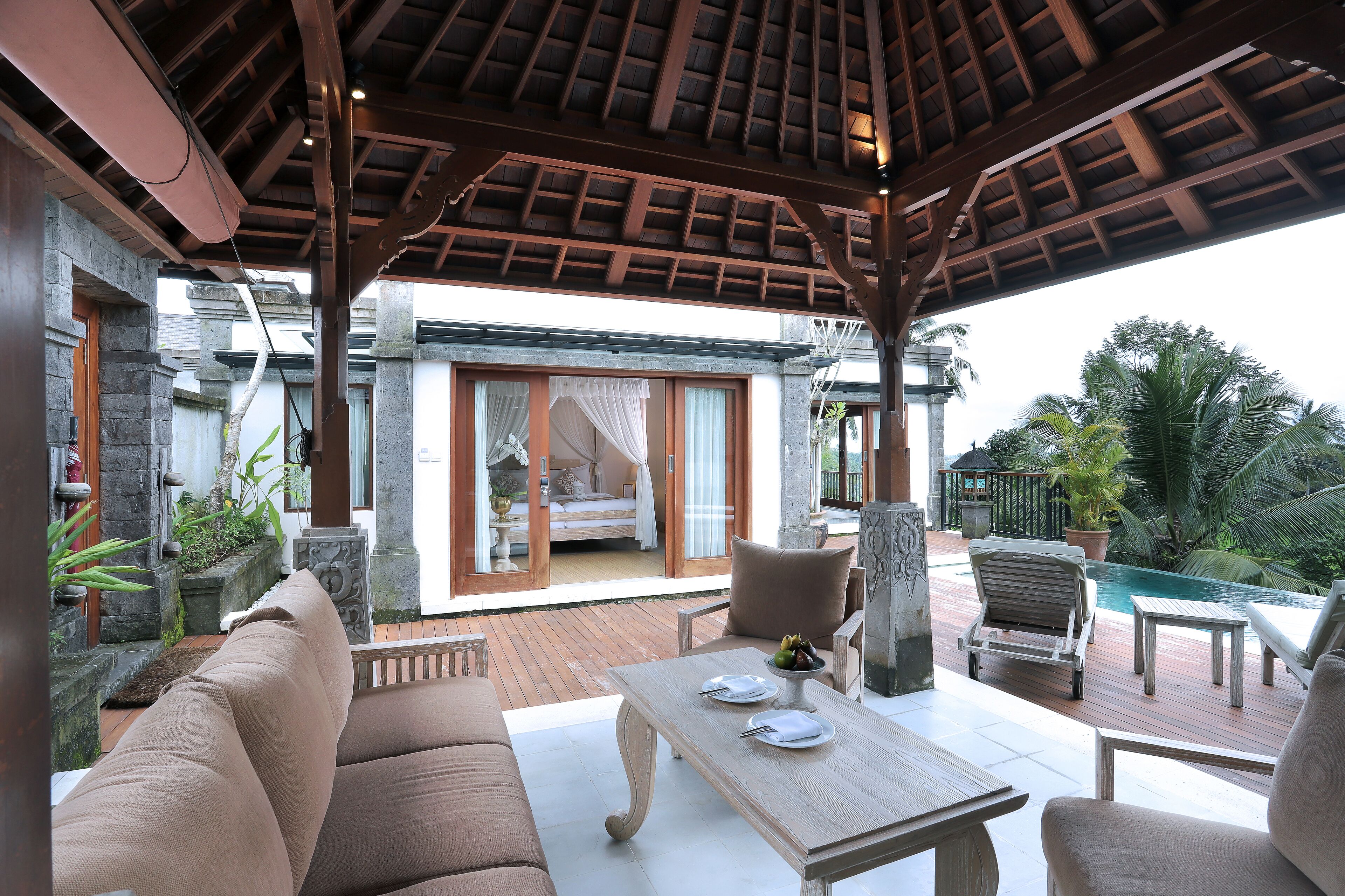 A covered outdoor seating area with a pool, wooden furniture, and a view of a bedroom with a bed and curtains.