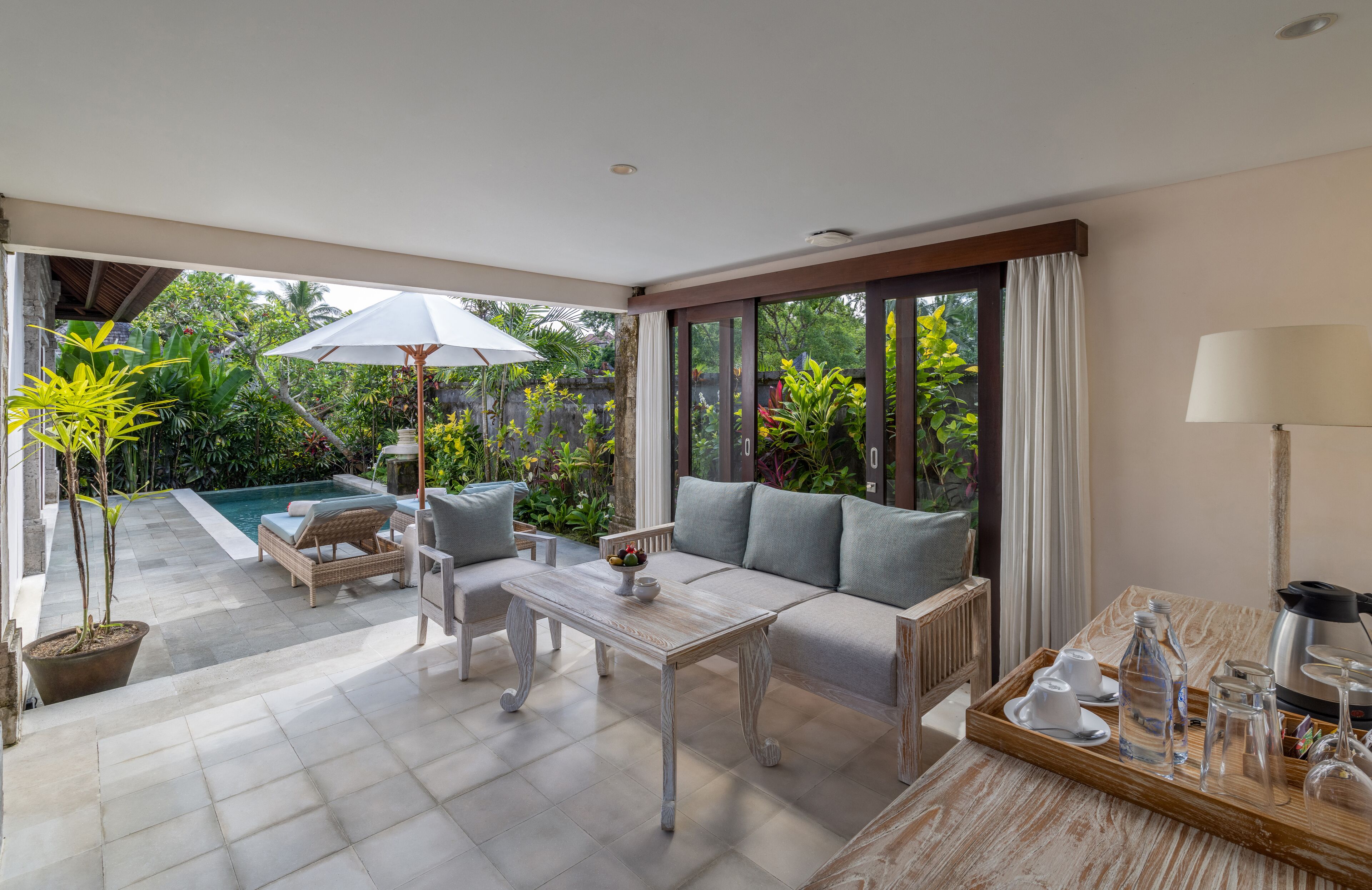 A modern room with a sofa, coffee table, and dining set. There is a large window with curtains, a potted plant, and a view of a pool.