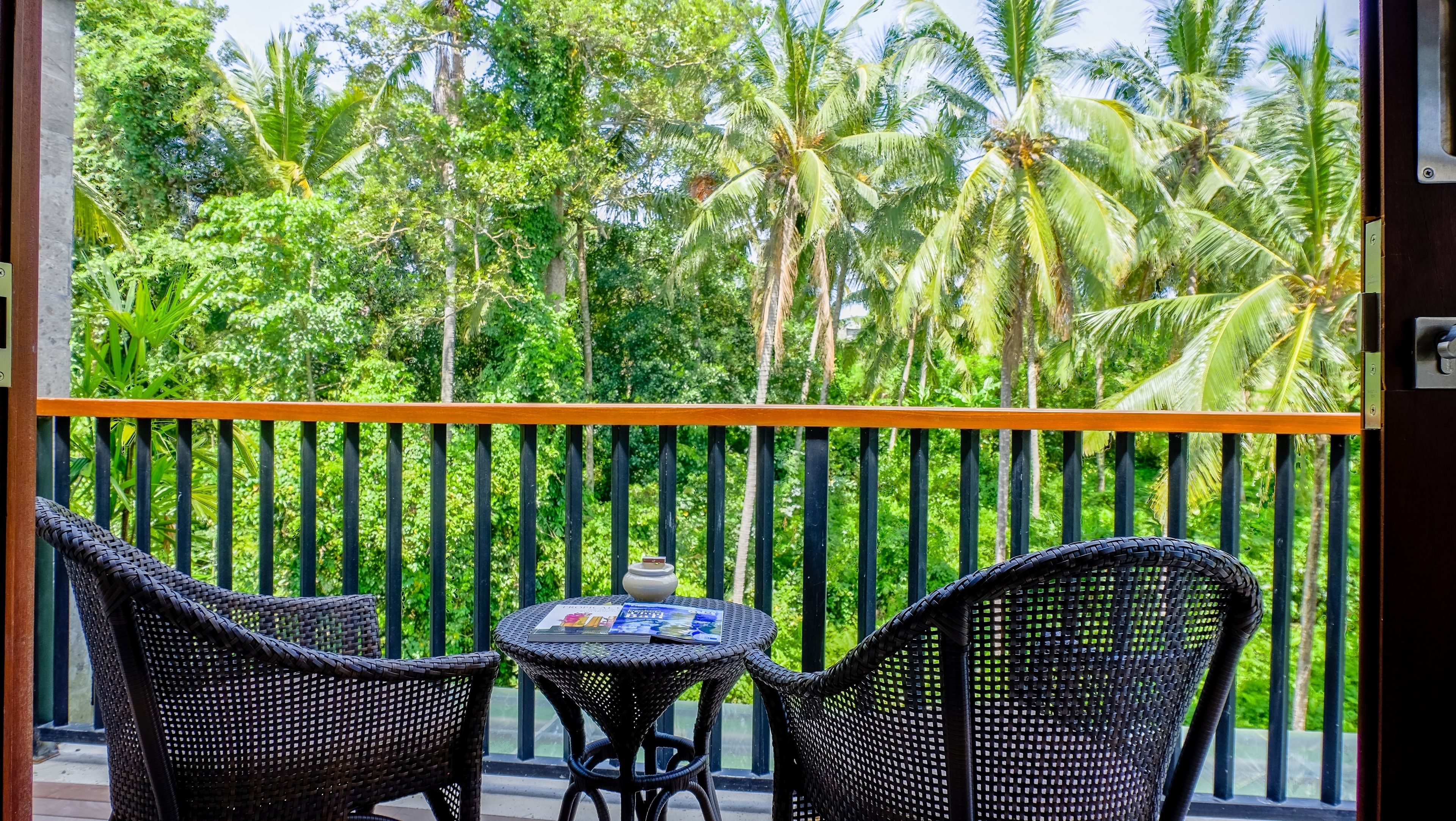 A balcony with wicker furniture, a small table, and a view of lush greenery and palm trees.