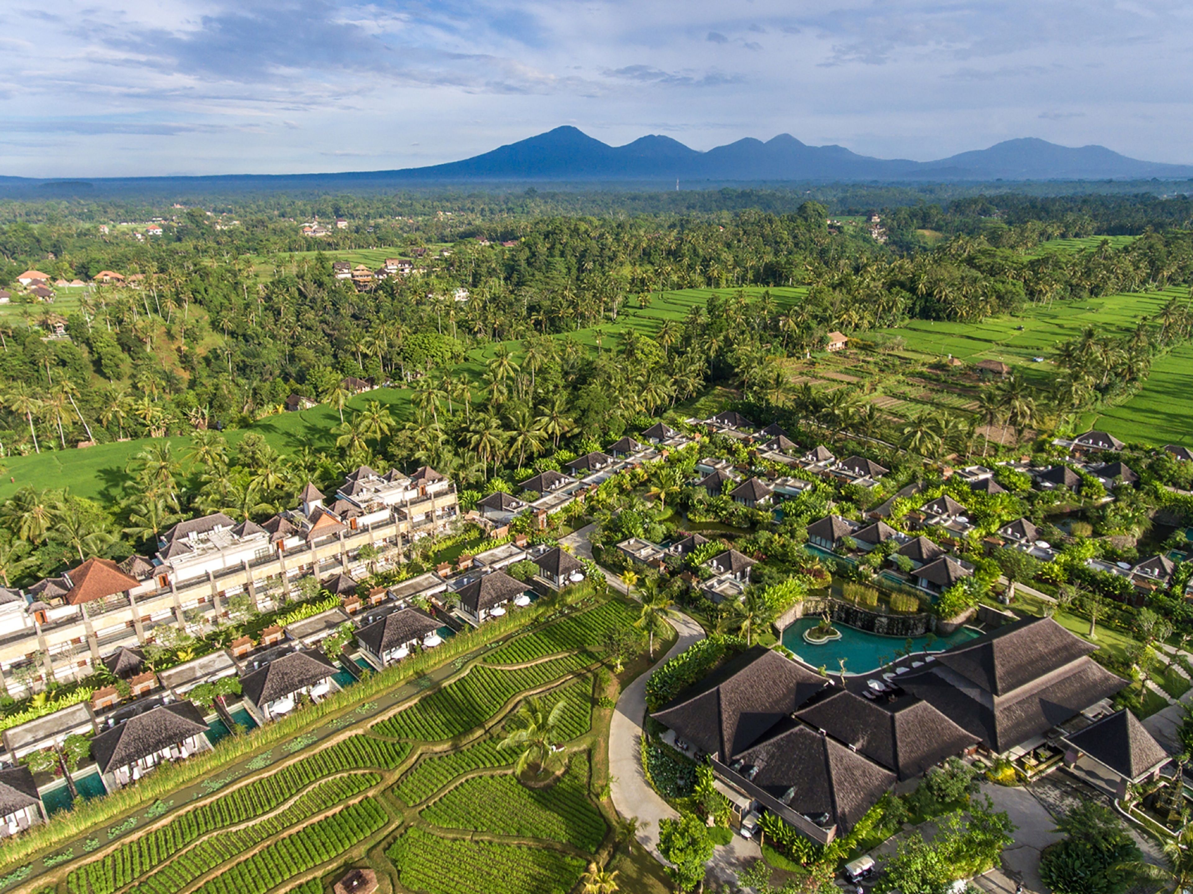 An aerial view of a resort with multiple buildings, including a large hotel with a swimming pool, surrounded by lush greenery and agricultural fields.