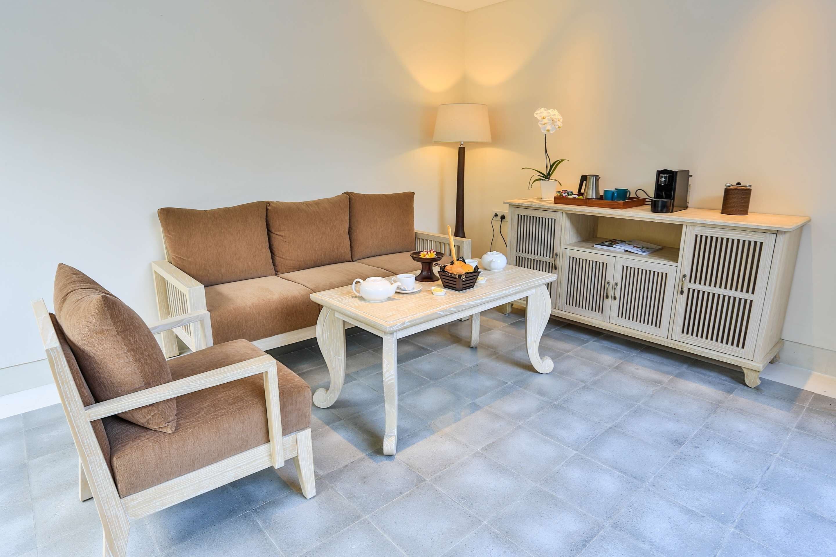 Un salón con un sofá color beige, un sillón de madera, una mesa de centro blanca, una lámpara, un mueble con una planta y una cafetera.