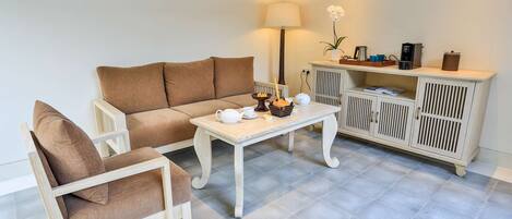 Un salón con un sofá color beige, un sillón de madera, una mesa de centro blanca, una lámpara, un mueble con una planta y una cafetera.