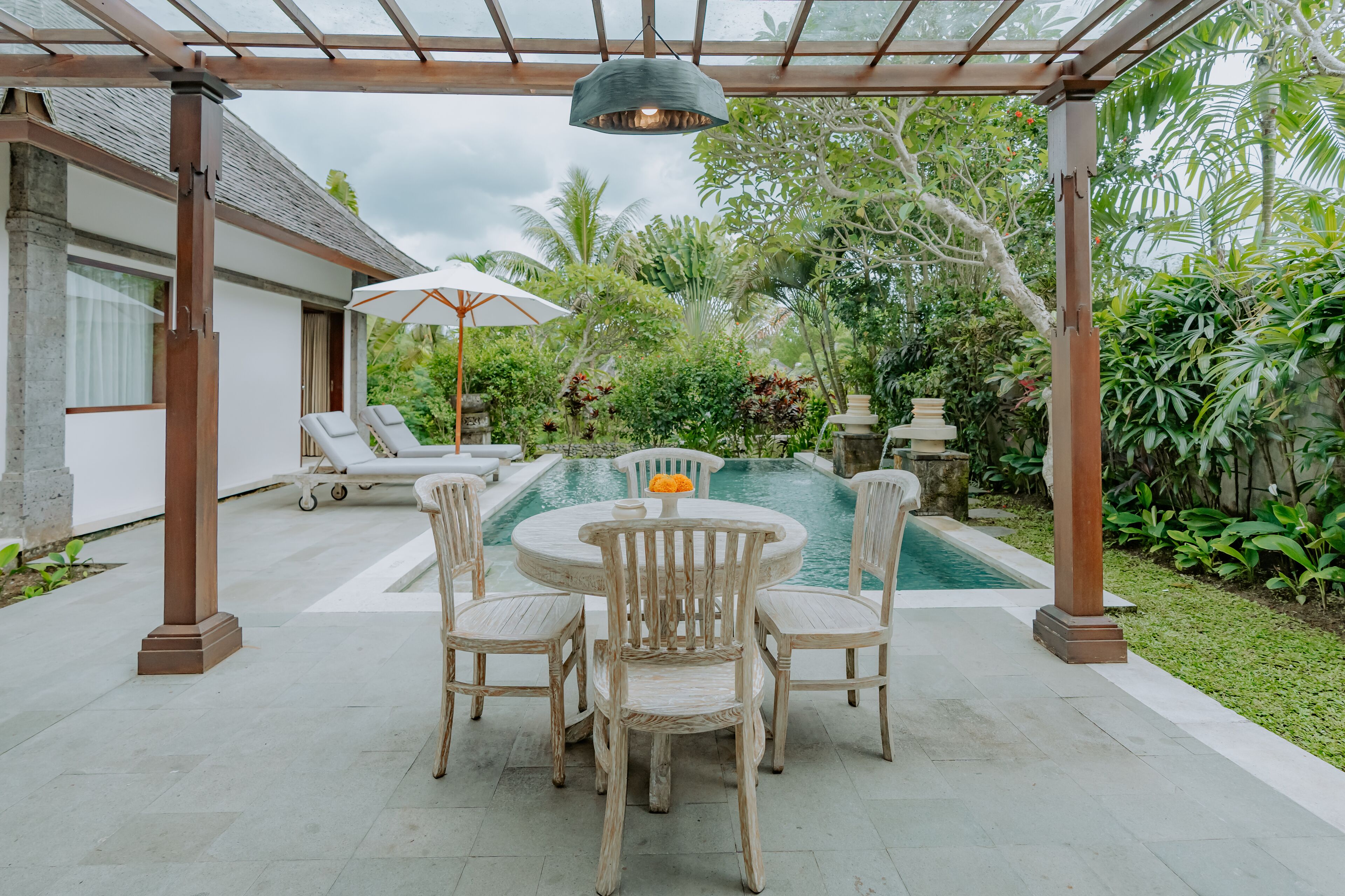 A covered outdoor area with a pool, lounge chairs, and a dining table with chairs.