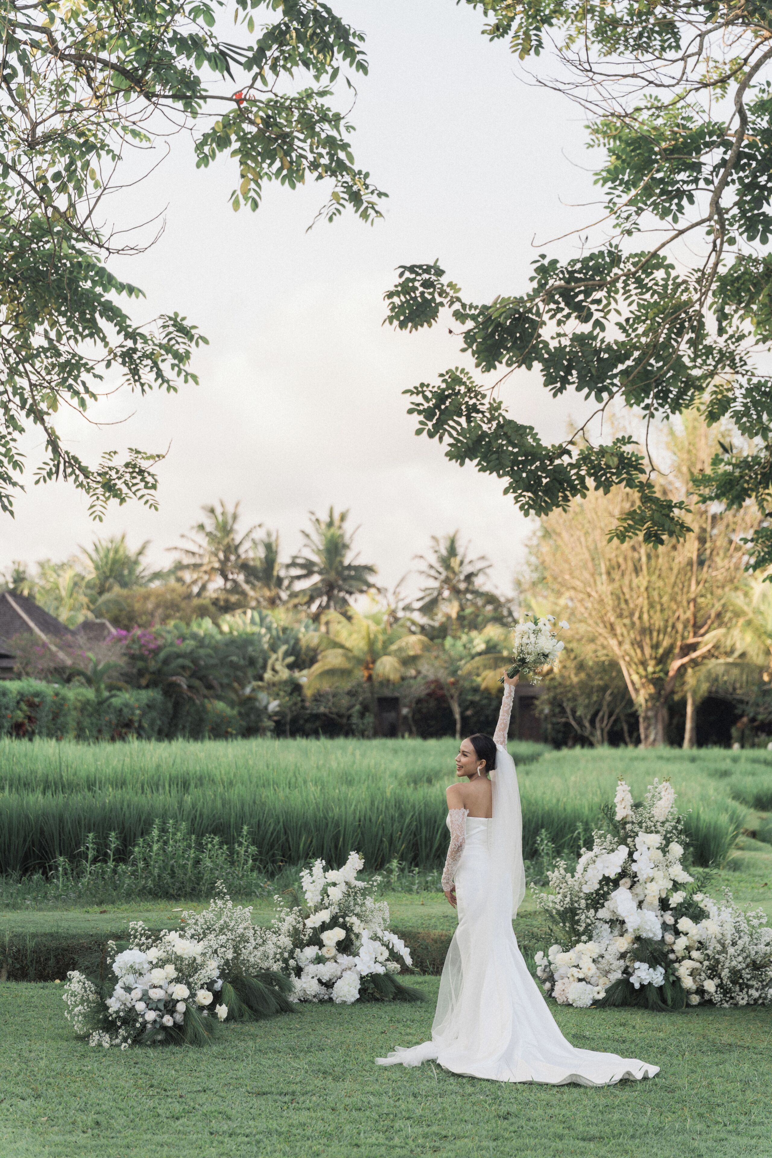 A bride in a white dress standing in a grassy area with white flowers and trees in the background.