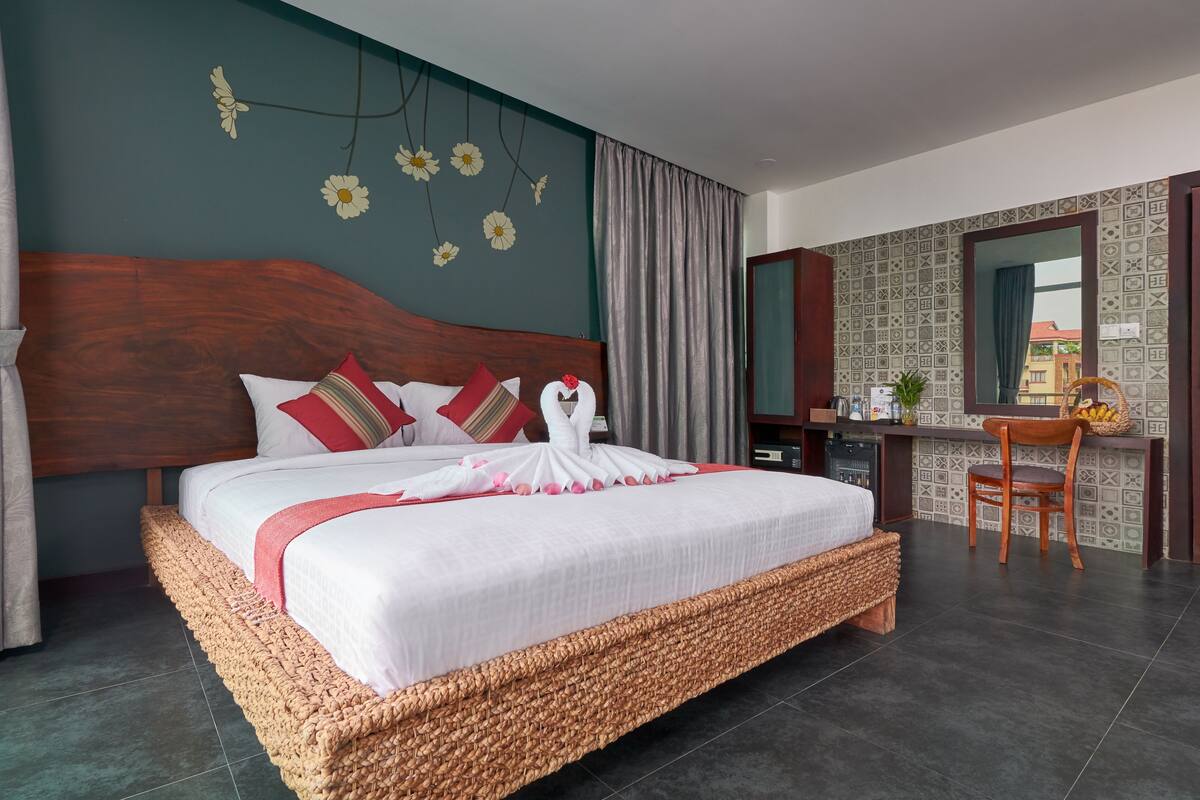 Una cama bien hecha con un almohadón decorativo con forma de cisne, un cabecero de mimbre, un escritorio con una silla y vistas de la ciudad a través de una ventana.