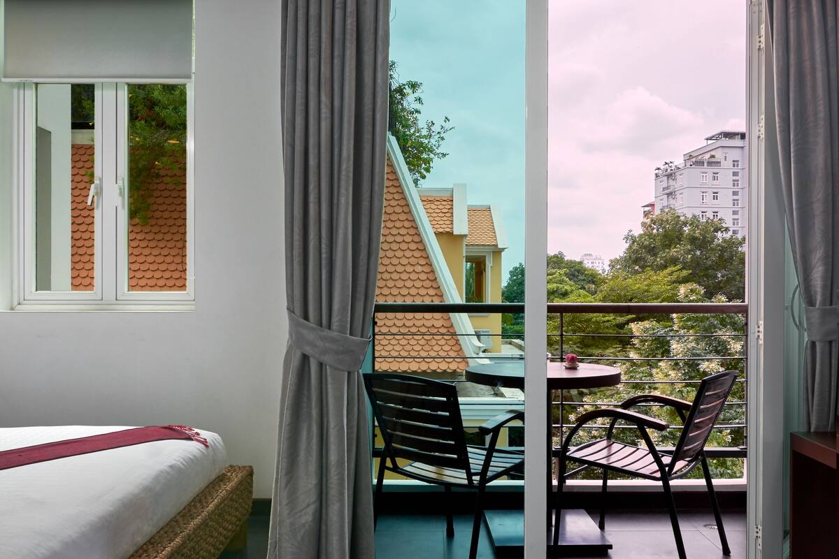 Una habitación con cama, un balcón con mesa y sillas, y vistas a edificios y árboles.