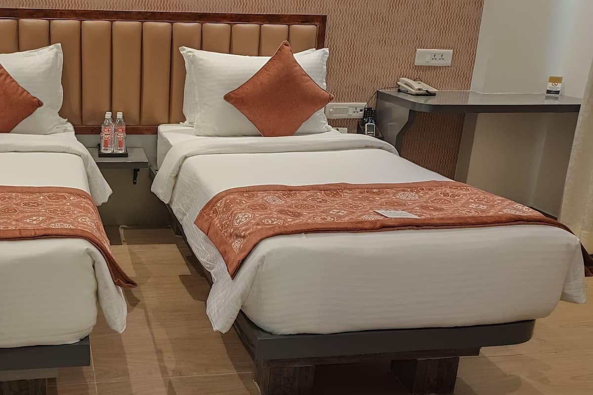 Ein Hotelzimmer mit zwei Einzelbetten, jeweils mit einer braunen, gemusterten Tagesdecke und weißen Kissen. Zwischen den Betten befindet sich ein kleiner Tisch mit einem Telefon und einer Wasserflasche.
