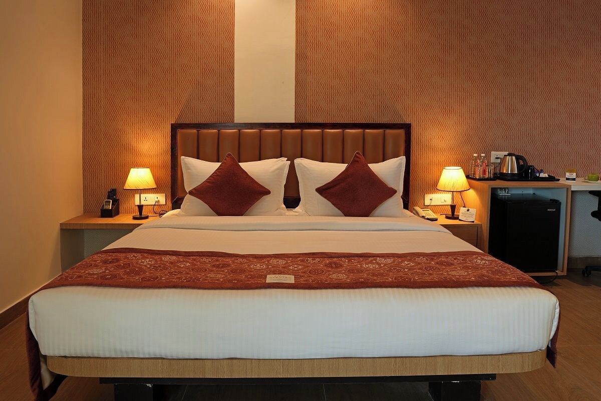 A hotel room with a large bed, two bedside lamps, a nightstand with a lamp, a small refrigerator, and a desk with a kettle and cups.