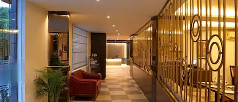 A hotel lobby with a checkered floor, a reception desk, a seating area with red chairs, and a decorative metal fence.