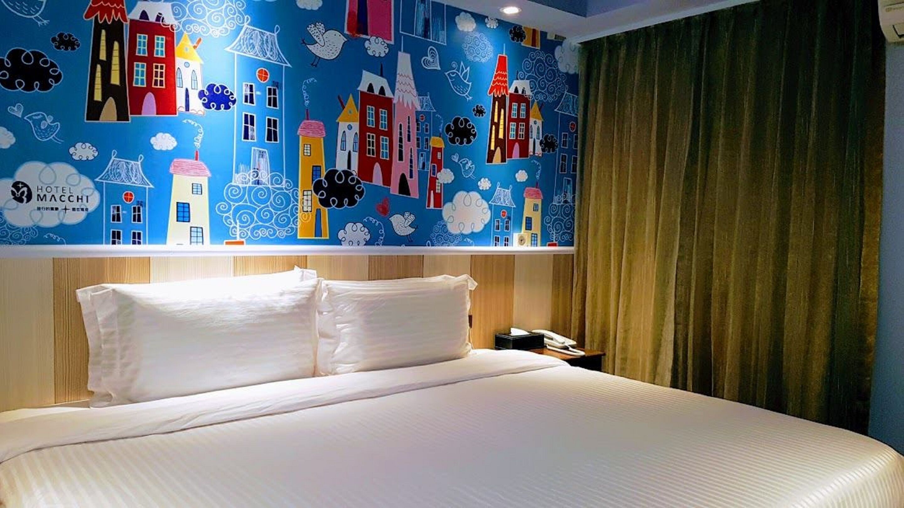 Ein Hotelzimmer mit Bett, Nachttisch und Wandgemälde mit bunten Gebäuden und Wolken.