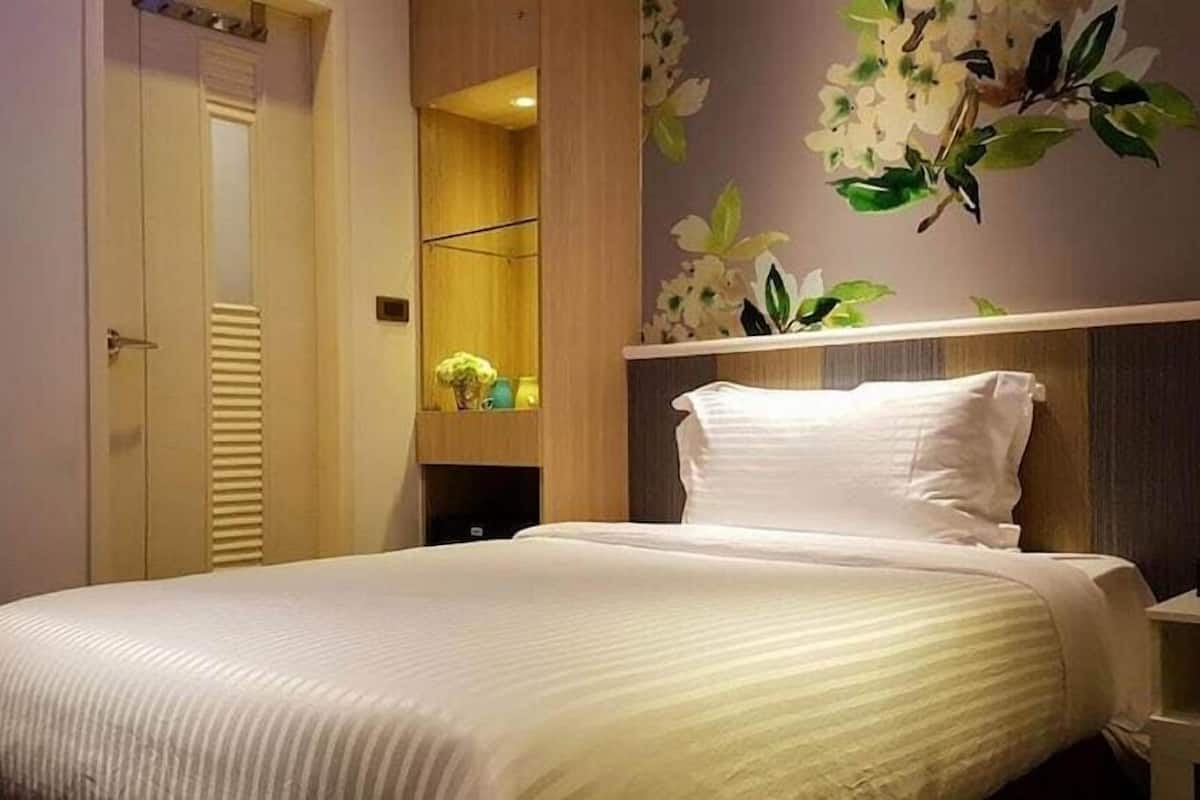 A neatly made bed with white linens, a wooden wardrobe, and a wall-mounted shelf with decorative items.