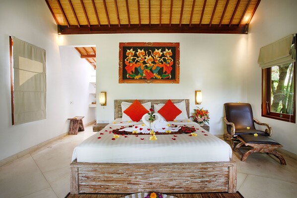 Habitación de hotel con una cama grande, cabecera de madera, un cuadro sobre la cama, una silla y ventana con vista a árboles.