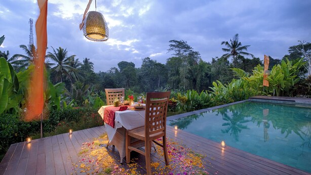 An outdoor dining area with a table set for two, surrounded by a wooden deck and a pool. - Villa Tegal Tis Ubud (Ubud)