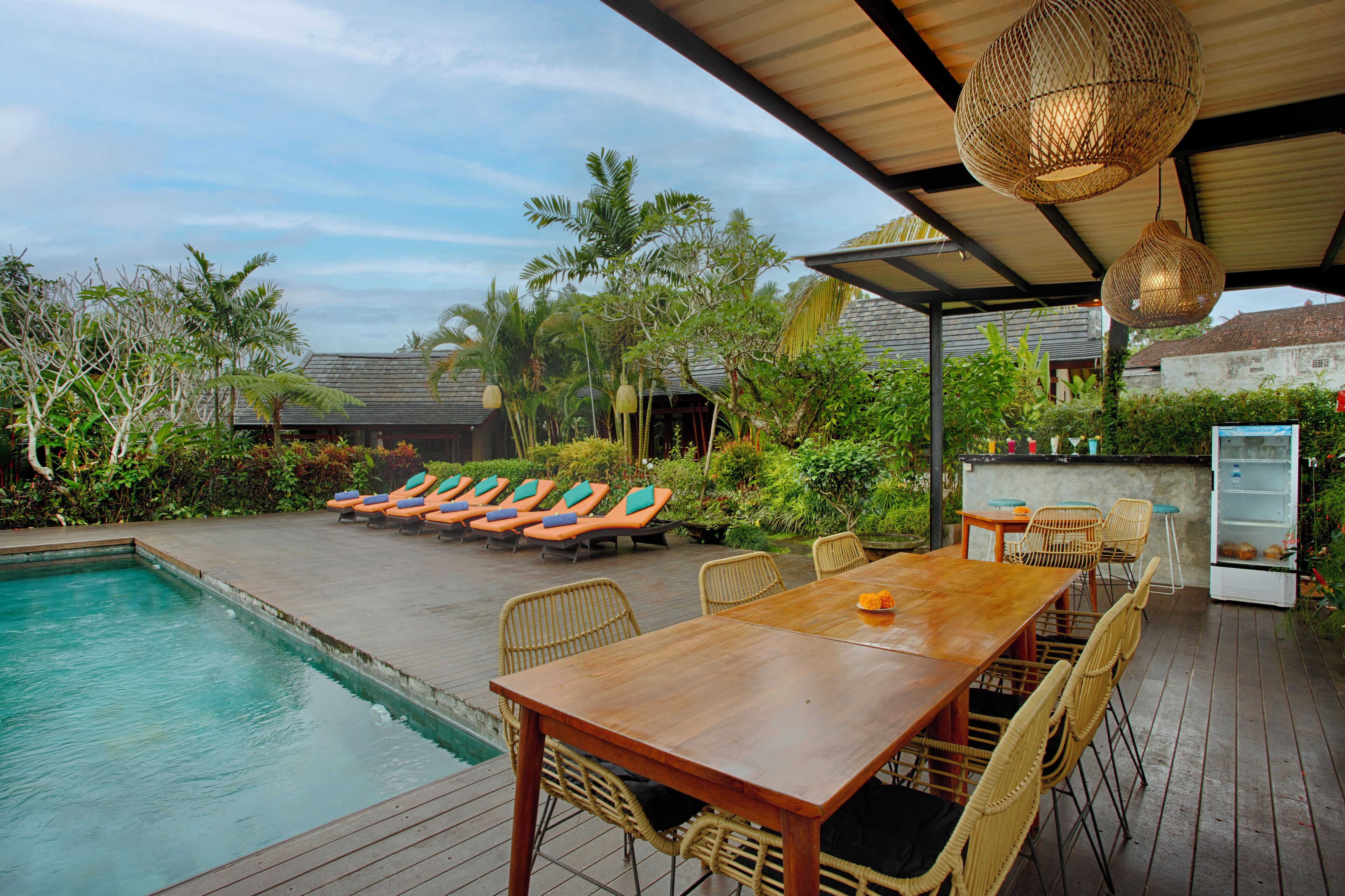 A poolside area with a wooden table and chairs, lounge chairs, and a covered seating area with hanging lights.