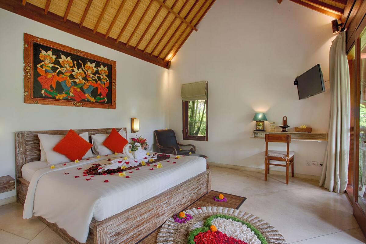Um quarto com uma cama grande, cabeceira de madeira, um quadro, uma cadeira, uma escrivaninha com abajur e uma janela com cortinas.