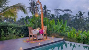 A poolside dining area with a table set for two, surrounded by lush greenery and a decorative hanging light. - Villa Tegal Tis Ubud (Ubud)